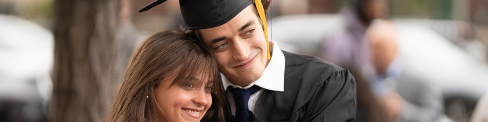 A student embracing a loved one outdoors after their graduation ceremony. They are wearing a cap and gown. Their loved one is wearing a red jacket. They are smiling for a photograph.