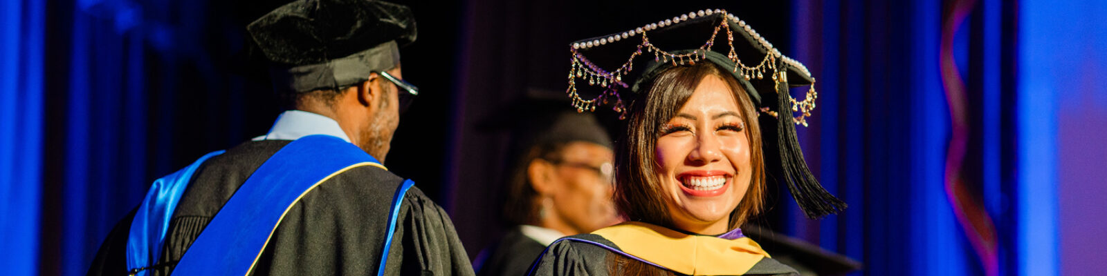 A student smiling widely while wearing regalia during their Graduate Hooding Ceremony